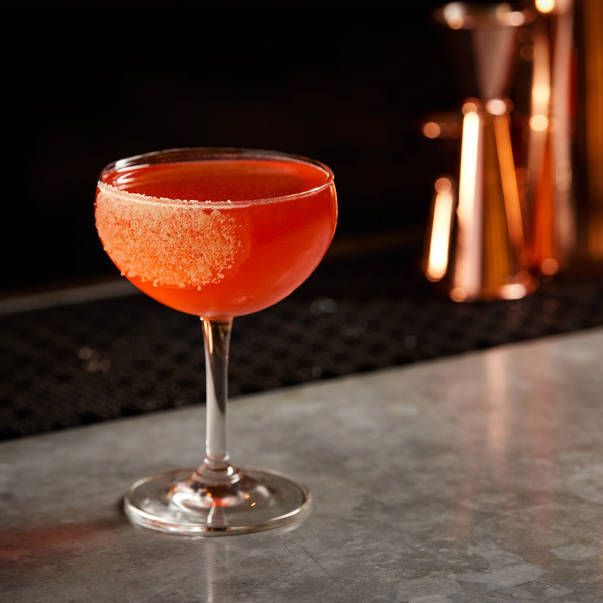 A salt-rimmed coupe with a deep red cocktail at a copper-lit bar.