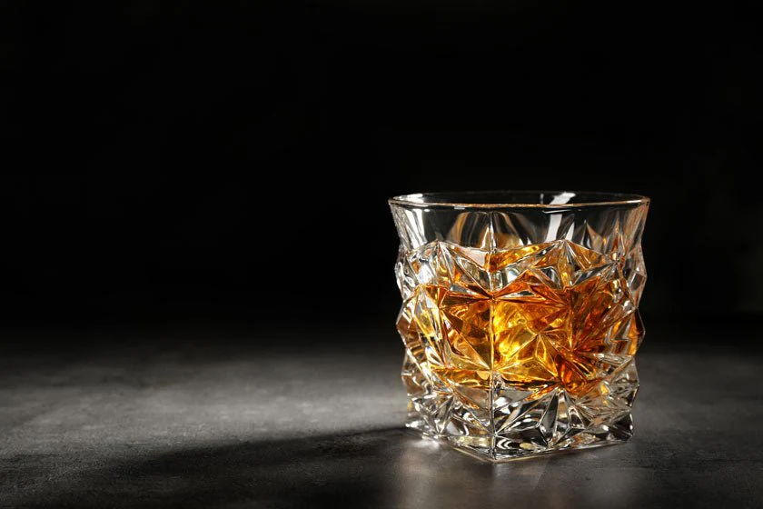 A heavy cut crystal rocks glass holding amber whiskey on a dark counter.